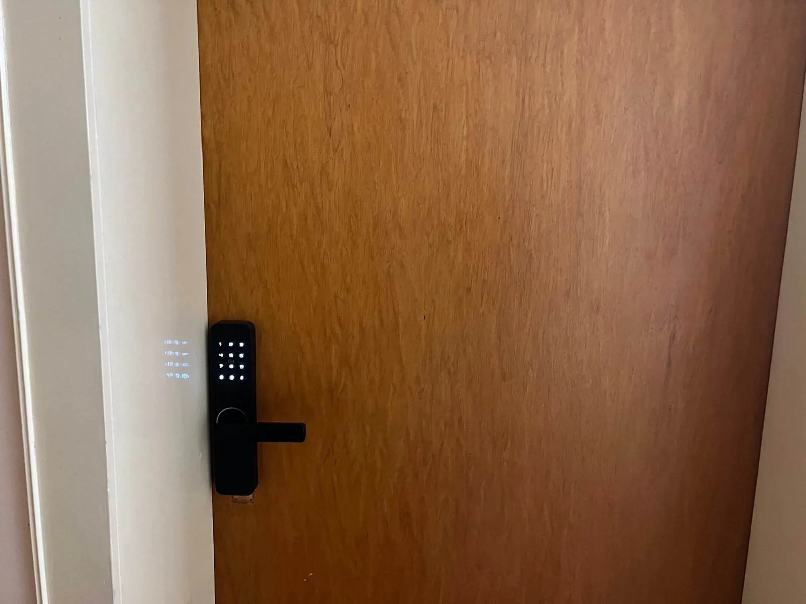 Abgebildet ist eine geschlossene Holztür mit einem digitalen Schloss und Touchscreen-Tastatur. Das Schloss ist schwarz, mit einer vertikalen Reihe beleuchteter Zahlen auf der linken Seite und einem horizontalen Türgriff. Die dezente Beleuchtung suggeriert einen modernen, sicheren Eingang.